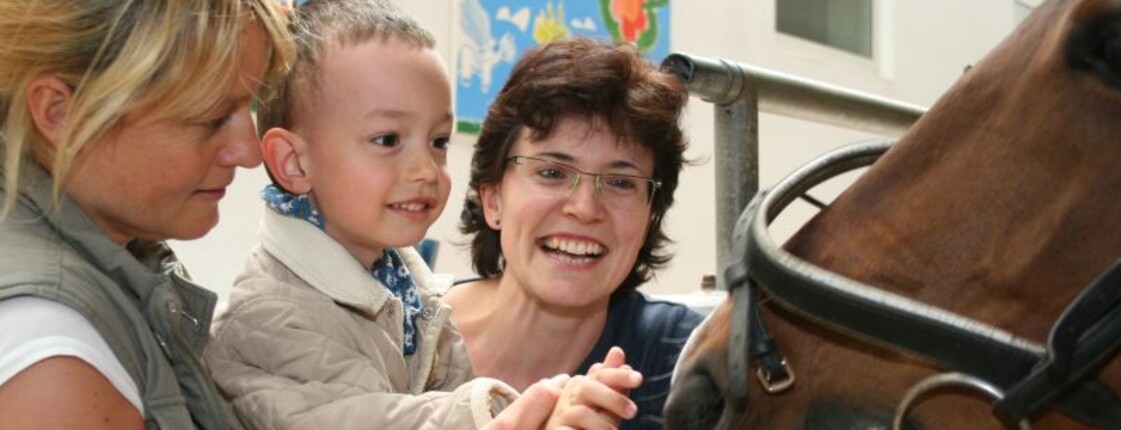 Integratives Reitzentrum Zwei Frauen führen einen kleinen Jungen mit seinem Arm an die Nüstern eines Pferdes heran.