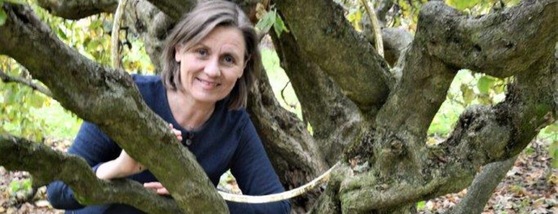 Frau mit halblangen Haaren hockt neben einem alten Baum.