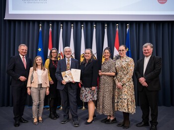 Journalistenpreisträger Martin Kranzl-Greinecker mit Edith Bürgler-Scheubmayr, Stellvertretende Direktorin der Caritas OÖ und dem UNSERE KINDER-Team sowie mit LH Thomas Stelzer (li) und Bischof Manfred Scheuer (re). Ein Gruppenfoto mit mehreren Menschen und in der Mitte steht der Preisträger mit einer Urkunde in der Hand.