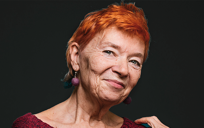 Das Porträt einer Frau ist zu sehen auf schwarzem Hintergrund. Es ist eine ältere Dame die lächelt, sie hat einen rothaarigen Kurzhaarschnitt. Links oben das Logo vom Caritas Wegweiser.
