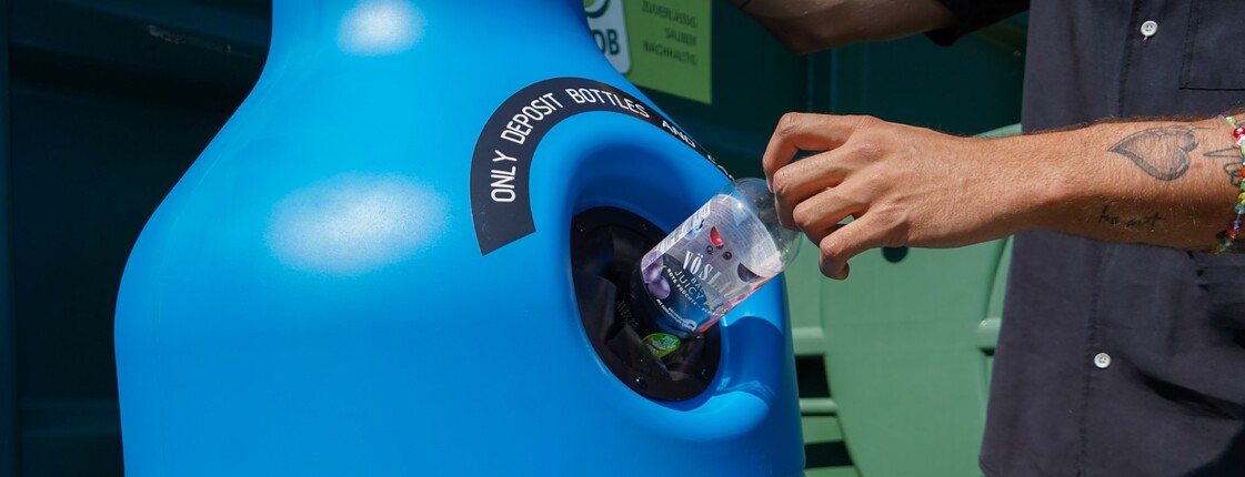Eine leere Pfandflasche wird in einen Pfandcontainer geworfen.