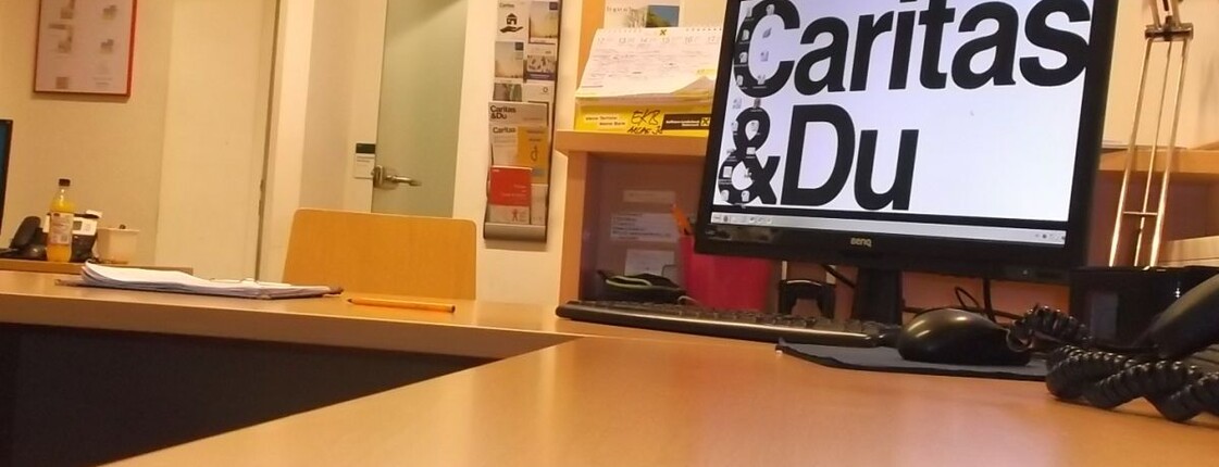 Arche 38 Büro Kontaktstelle Schreibtisch mit Computer und als Desktop-Hintergrund der Schriftzug "Caritas&Du"