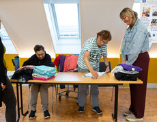 Zwei Frauen legen Wäsche zusammen, die auf einem Tisch liegt. Zwei weitere Frauen sehen ihnen dabei zu. Im Hintergrund ist ein Plakat mit "Wäscherei St. Pius" zu sehen.