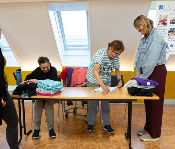 Zwei Frauen legen Wäsche zusammen, die auf einem Tisch liegt. Zwei weitere Frauen sehen ihnen dabei zu. Im Hintergrund ist ein Plakat mit "Wäscherei St. Pius" zu sehen.