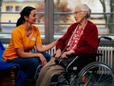Links eine junge Frau in orangem Shirt sitzt und hält den Arm einer alten Dame, die im Rollstuhl sitzt.