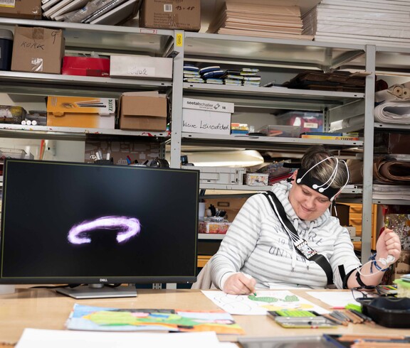 Frau zeichnet in einem Atelier ein Bild und ist von technischen Geräten umgeben.