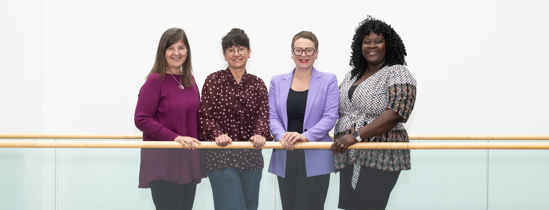 Vier Frauen stehen vor einem Geländer aus Glas in einem Gebäude zusammen.