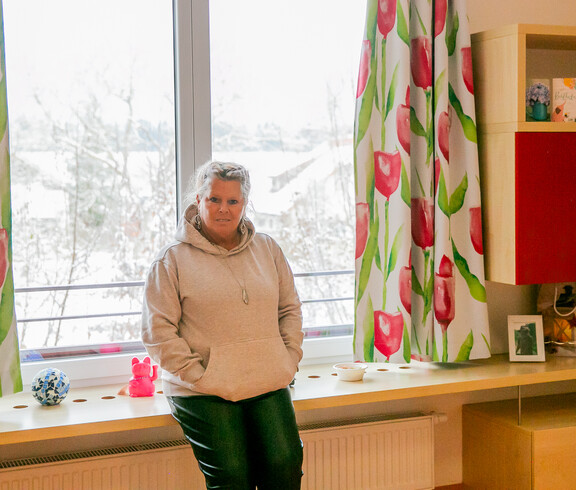 Eine Frau in beigem Hoodie und dunkler Hose steht vor einem Fenster in iherm Zimmer und sieht in die Kamera.
