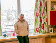 Eine Frau in beigem Hoodie und dunkler Hose steht vor einem Fenster in iherm Zimmer und sieht in die Kamera.