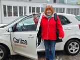 Eine Frau mit roter Jacke steht vor einem Caritas Auto mit offener Fahrertür