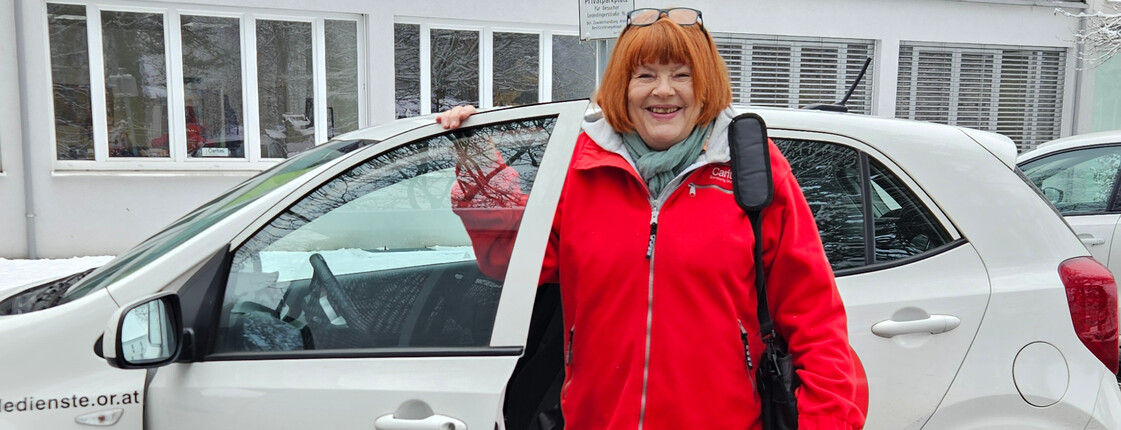 Eine Frau mit roter Jacke steht vor einem Caritas Auto mit offener Fahrertür