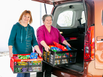 Ulrike Mayer (Caritas-Mitarbeiterin) und Maria Huber Zwei Frauen stehen bei einem Auto mit offenen Kofferraum und halten Kisten mit Lebensmittel in der Hand.