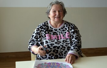 eine Frau mit kürzeren grauen Haaren sitzt an einem Tisch mit einer Schere in der Hand.