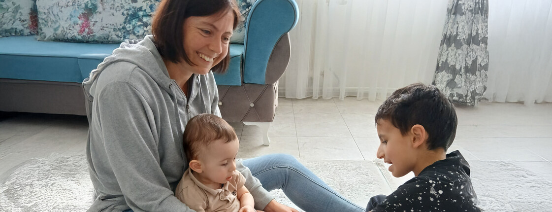 Eine Frau sitzt am Boden und spielt mit zwei Buben, die ebenefalls am Boden sitzten.