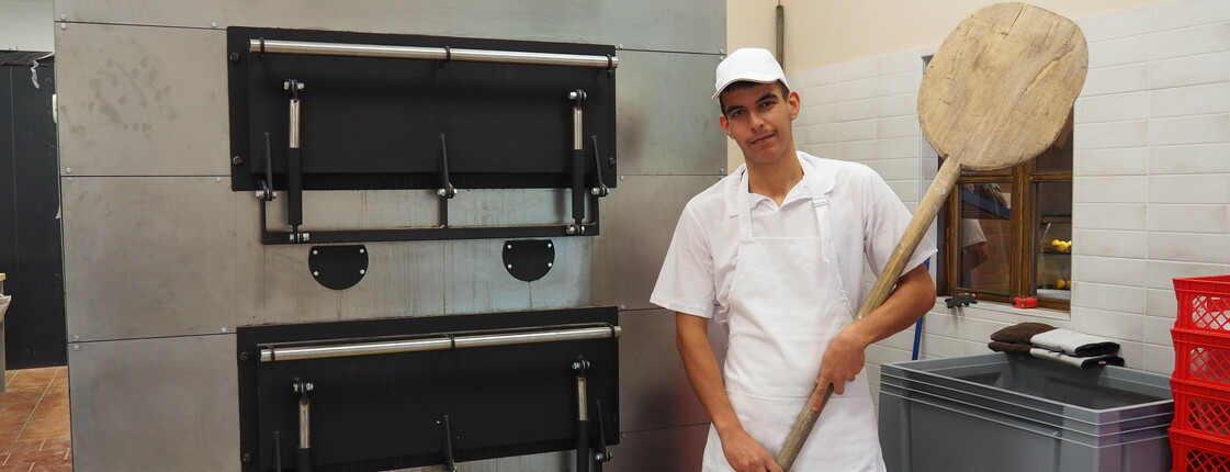 Ein junger Mann mit Brotschieber rechts vor einem großen Backofen stehend