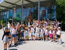 Kinder stehen für ein Gurppenfoto vor dem Zoo Eingang und haben die Hände in die Luft gestreckt.