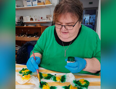 Frau in grünem T-Shirt und blauen Handschuhen bemalt einen Seidenschal mit Sonnenblumen.