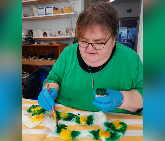 Frau in grünem T-Shirt und blauen Handschuhen bemalt einen Seidenschal mit Sonnenblumen.