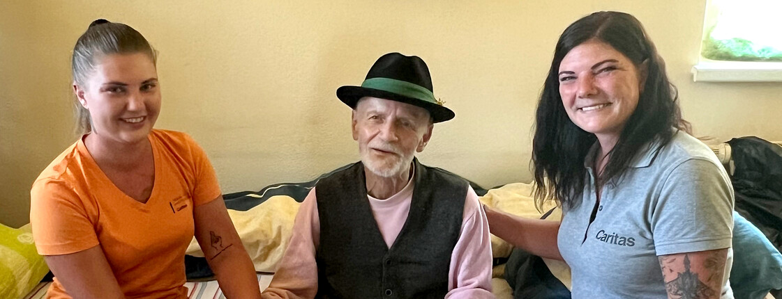 Zwei Caritas-Pflegerinnen sitzen neben einem alten Mann mit Steirer-Hut.