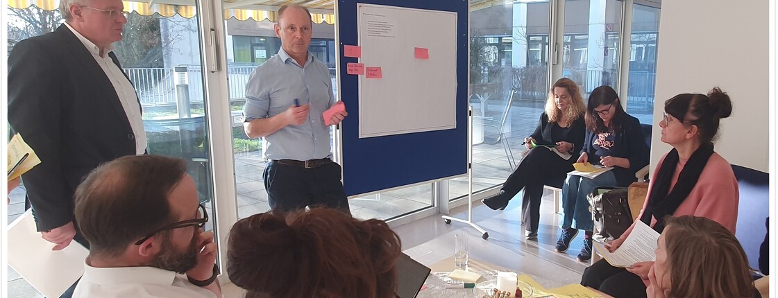 Mehrere Erwachsene sitzen um eine Pinnwandtafel. Typische Workshop Situation.