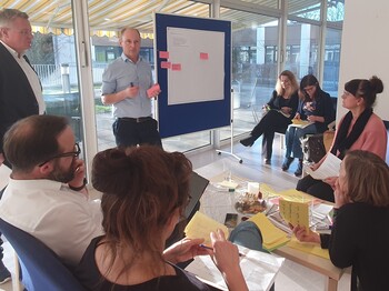 Mehrere Erwachsene sitzen um eine Pinnwandtafel. Typische Workshop Situation.