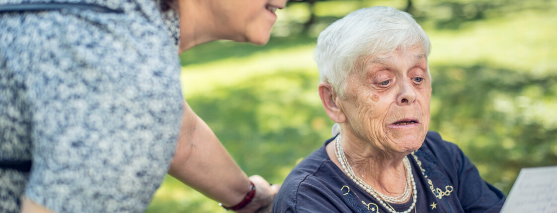 Eine Seniorin sitzt in einem Korbstuhl in einem Garten und liest eine Postkarte, welche sie in der Hand hochhält. Eine Pflegerin steht neben ihr und schaut ebenfalls die Postkarte an.