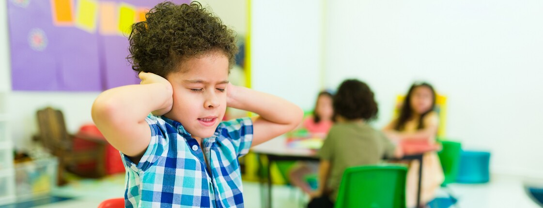 Kind sitzt an einem Tisch in einer Schulklasse und hält sich die Ohren zu.