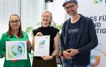 Vier Personen stehen und schauen in die Kamera. die beiden Frauen in der Mitte halten jeweils ein kleines Bild mit Klimabündnispartner Logo in der Hand.