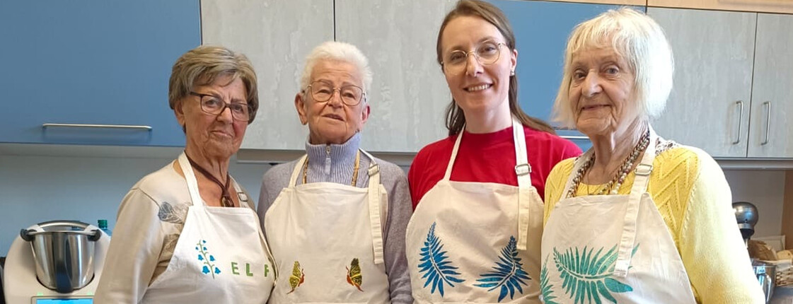 Drei ältere Damen und eine junge Frau stehen mit umgebundenen Kochschürzen in der Küche.