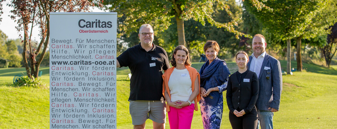 Zwei Männer und drei Frauen stehen neben einem Caritas Schild auf einer Wiese. Im Hintergrund sind Bäume.