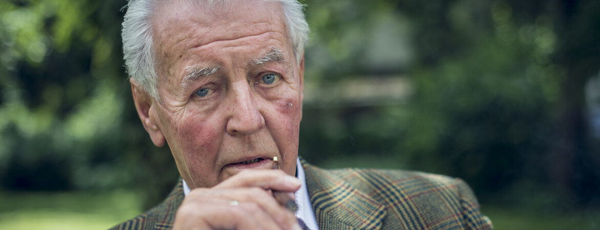 Portrait eines älteren Herrn, der in einem Garten sitzt und raucht.