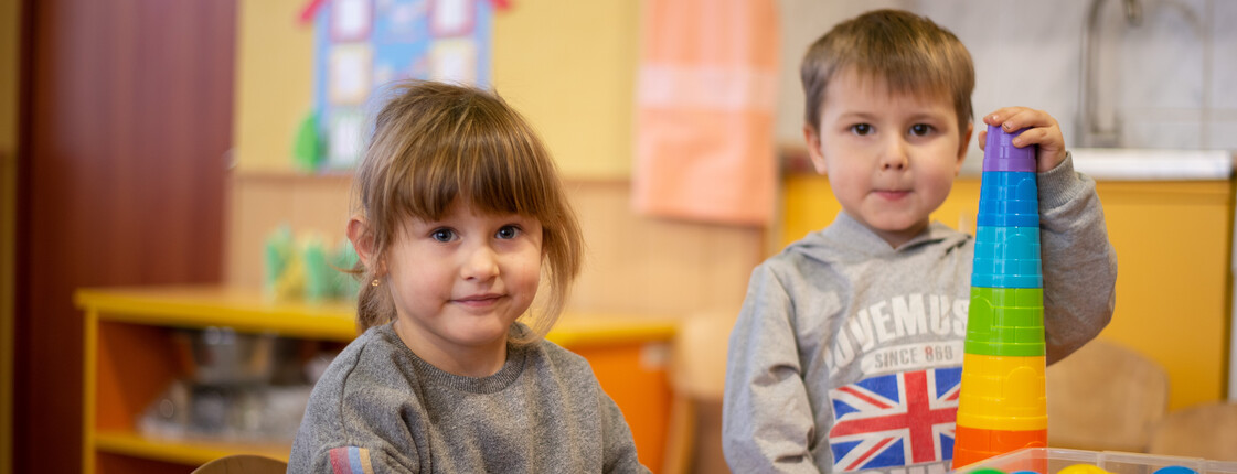 Zwei Kinder spielen im Kinder- und Familienzentrum Charkiw, Ukraine. Ein Junge baut einen Turm aus bunten Bechern, während ein Mädchen lächelt.