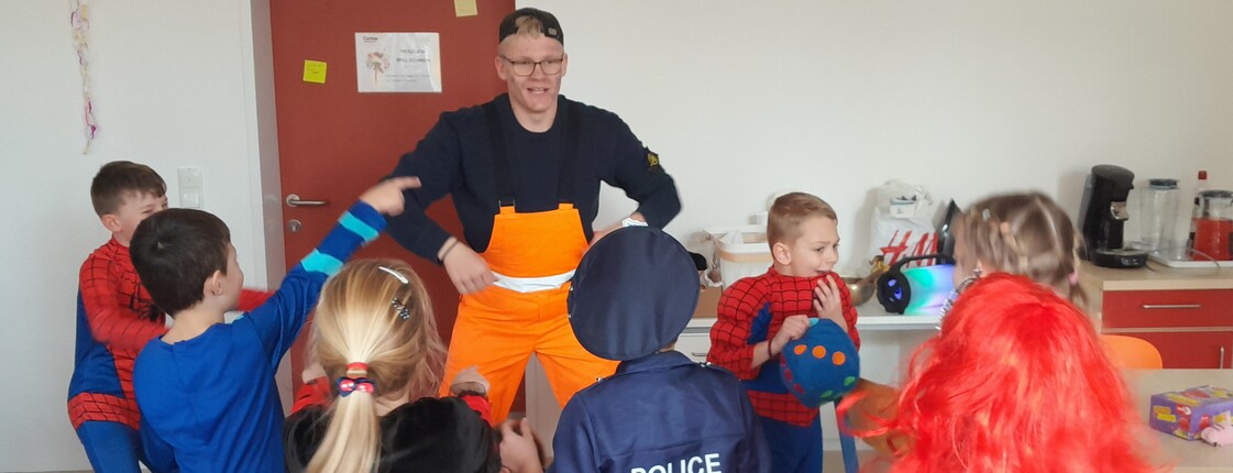 Junger Mann in gelber Latzhose ist umzingelt von mehreren Kindern in verschiedenen Kostümen.