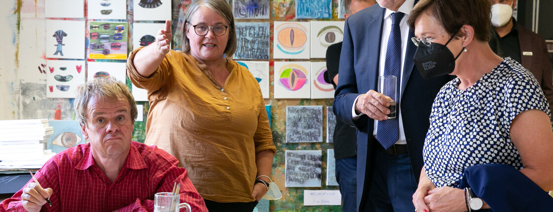Vier Personen in der Kunstwerkstätte St. Pius. Rechts Bundespräsident Van der Bellen und seine Gattin, stehend neben einem Tisch mit Zeichungen.