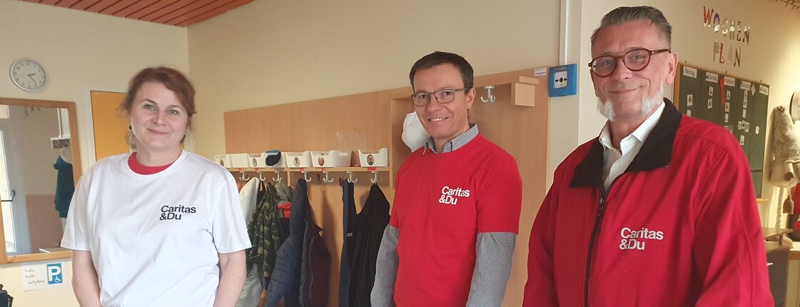 Drei Erwachsene stehen vor einem Tischfussballtisch, eine Frau im weißen T-Shirt ganz links und rechts zwei Männer mit roter Oberbekleidung.