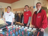 Drei Erwachsene stehen vor einem Tischfussballtisch, eine Frau im weißen T-Shirt ganz links und rechts zwei Männer mit roter Oberbekleidung.
