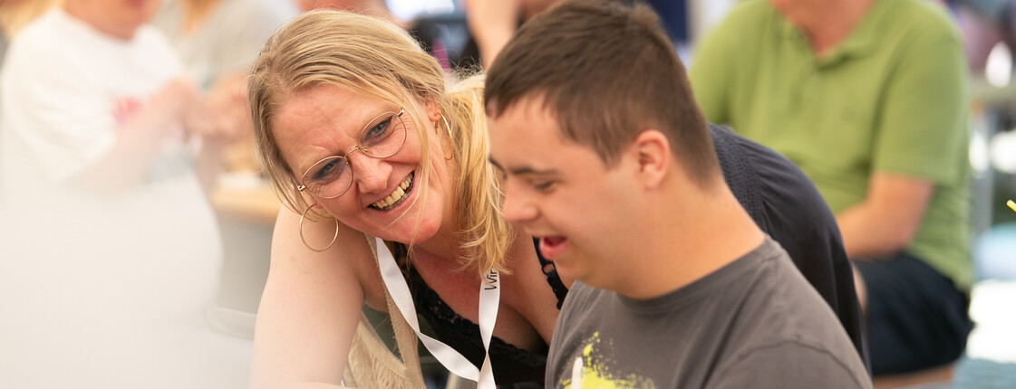 Eine Betreuerin beugt sich zu einem Menschen mit Beeinträchtigungen und lächelt ihn an.