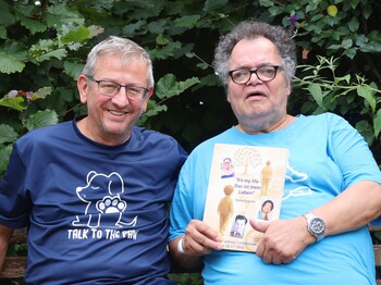 Zwei ältere Männer sitzen auf einer Bank und schauen in die Kamera. Der Mann mit Brille rechts hält ein Buch in der Hand.