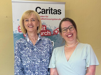 Mag.a Birgit Ippisch (li.) mit ihrer Nachfolgerin Sandra Schwaiger. Zwei Frauen stehen vor einem Caritas Roll-Up und lächeln.