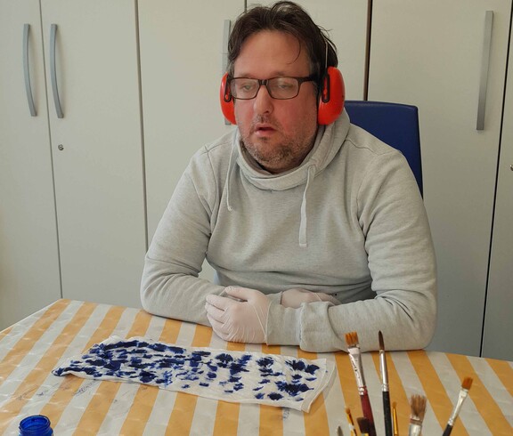 Mann im weißen Pulli mit Kopfhörer, vor ihm liegt ein Stück bemalter Seidenstoff