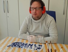 Mann im weißen Pulli mit Kopfhörer, vor ihm liegt ein Stück bemalter Seidenstoff