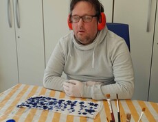 Mann im weißen Pulli mit Kopfhörer, vor ihm liegt ein Stück bemalter Seidenstoff