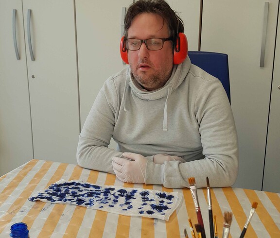 Mann im weißen Pulli mit Kopfhörer, vor ihm liegt ein Stück bemalter Seidenstoff