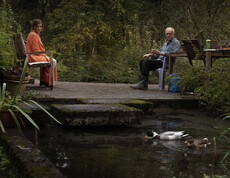 Frau und Mann sitzen im Garten, davor schwimmt eine Ente im Teich