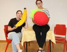 Zwei Frauen mit überdimensionierten Früchten in Händen, eine sitzt am Tisch