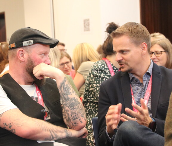 Zwei Männer sitzen in einem gefüllten Saal und unterhalten sich.