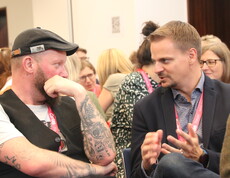 Zwei Männer sitzen in einem gefüllten Saal und unterhalten sich.