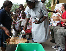 Krankenschwester gießt heißes Wasser in einen großen Topf mit Brei, umgeben von Kindern und einer weiteren Helferin in der Ernährungsstation in N’sele