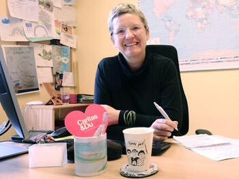 Frau sitzt vor Landkarte am Computer, dafür ein feministisches Häferl und ein Caritas&Du-Herz