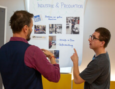 Zwei Männer stehen vor einem Plakat und sprechen miteinander. Auf dem Plakat steht "Unsere Kunden in der Industrie und Produktion."